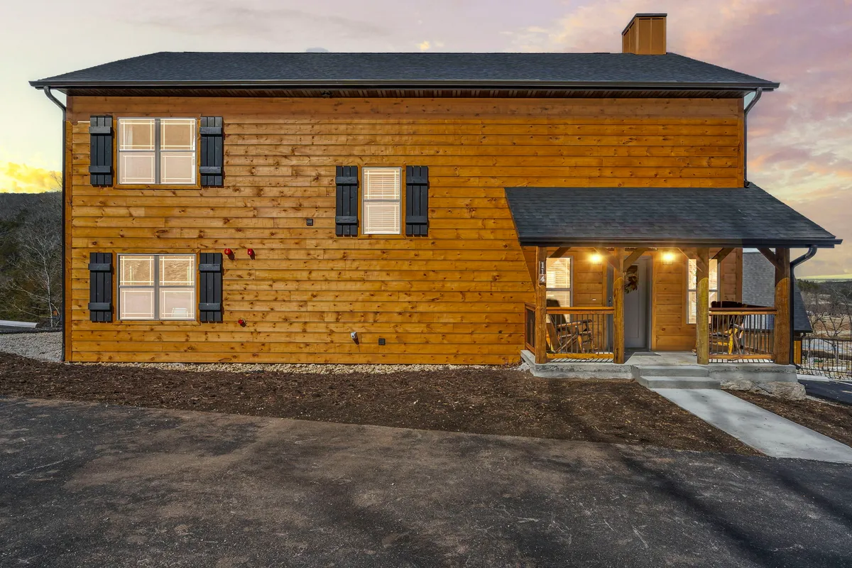 A cozy exterior shot of a cabin's wooden exterior with shuttered windows and a softly glowing porch light. The porch area is inviting, set up for relaxation and surrounded by nature, suggesting a tranquil spot to unwind after adventures or enjoy quiet moments with a pet.