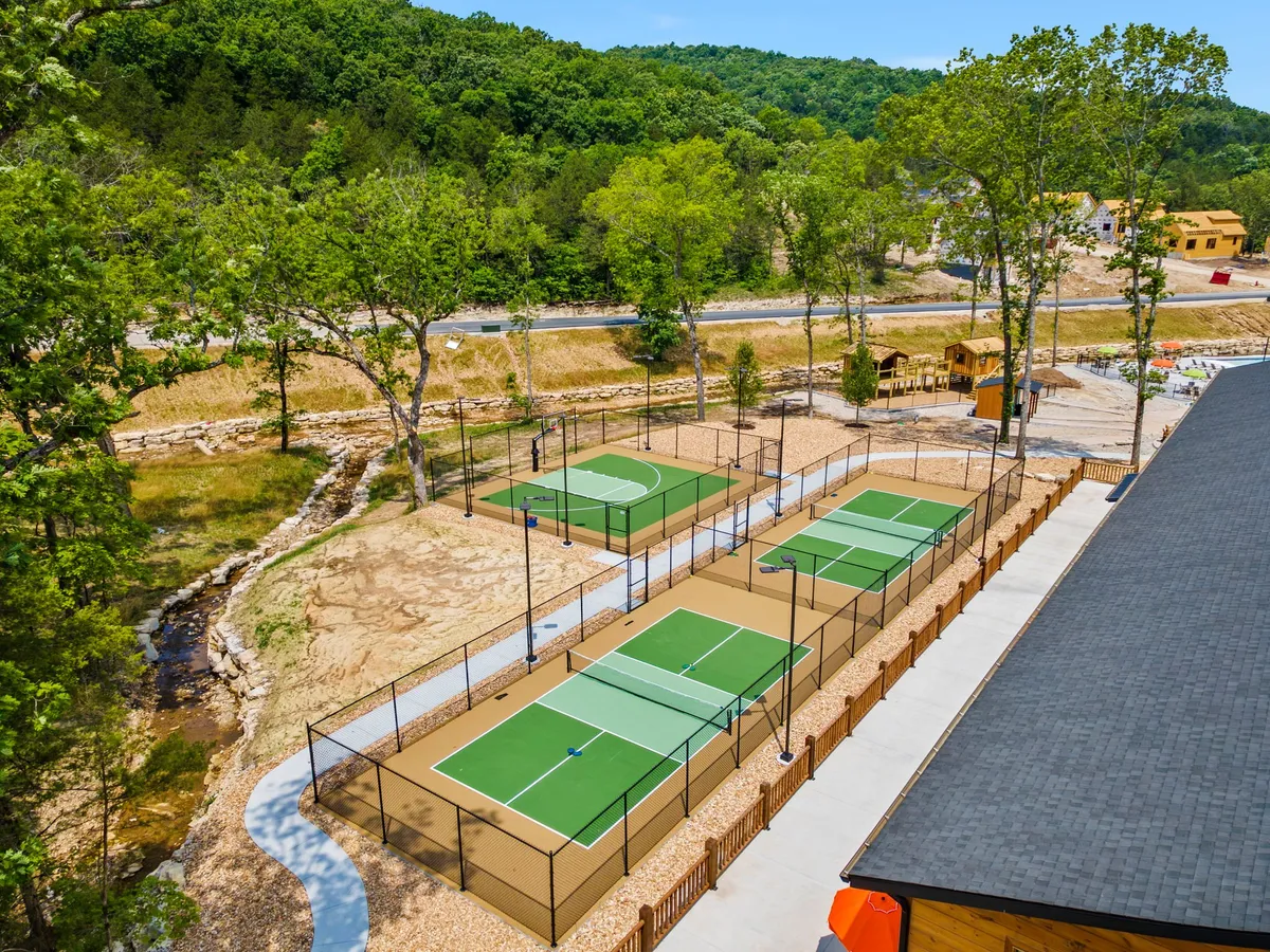 A bright exterior image showing tennis courts set amidst greenery, with a visible playground area nearby. Families and children can be seen enjoying the amenities, highlighting activity and togetherness.