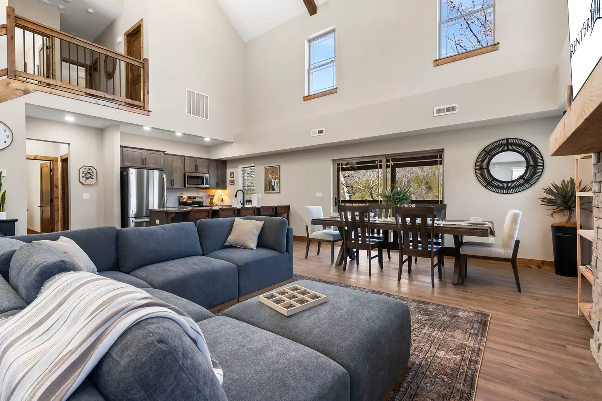 This image features a welcoming open-concept space that seamlessly connects the living room, dining area, and kitchen. The spacious sofa faces a dining table, while the kitchen area with large windows and wooden beams is visible in the background. Natural light bathes the entire area, enhancing the inviting, communal atmosphere.