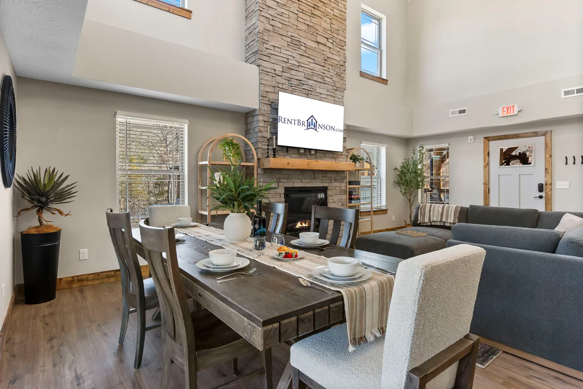 The image showcases the cozy interior of the cabin, featuring a spacious dining table set for gatherings, a glowing fireplace, a comfortable sofa, and a large television—perfect for family meals, relaxing evenings, and entertaining. The warm lighting and inviting decor create a welcoming atmosphere ideal for memorable getaways.
