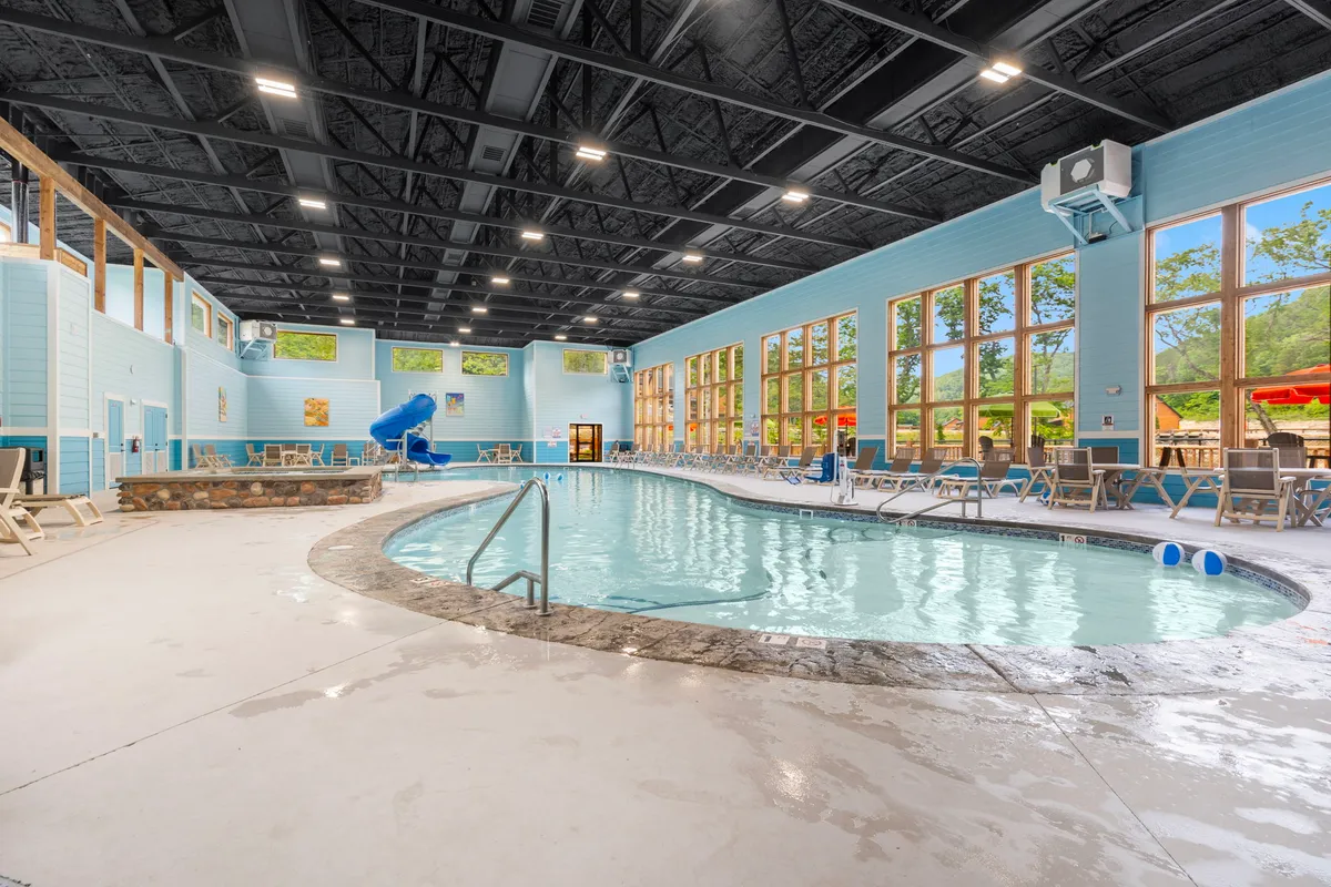A vibrant indoor swimming pool featuring a blue water slide, large windows letting in natural light, and several lounge chairs arranged for relaxing. The atmosphere is bright and welcoming, perfect for family gatherings or letting kids play safely indoors.