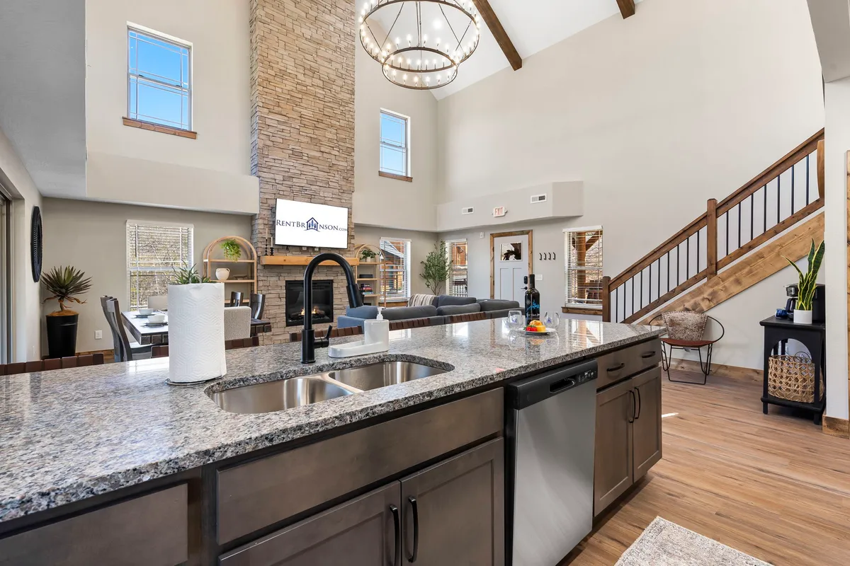 A beautifully designed open-plan room showcasing a modern kitchen with granite countertops and high-end appliances, a stone fireplace, and a spacious living area with large windows and wooden beams. The setting is elegant yet inviting, encouraging both cooking and socializing.
