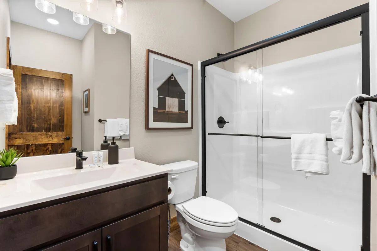 A modern and inviting bathroom featuring a vanity with sink, framed wall art, a glass shower enclosure, and a wooden door. The space feels fresh and functional, designed for both relaxation and convenience.
