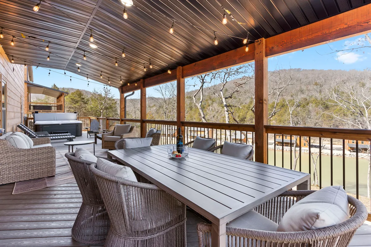 An outdoor dining table with woven chairs set on a deck, complemented by string lights and a hot tub. The setting boasts a beautiful mountain backdrop, creating a serene environment for outdoor meals and evening relaxation.