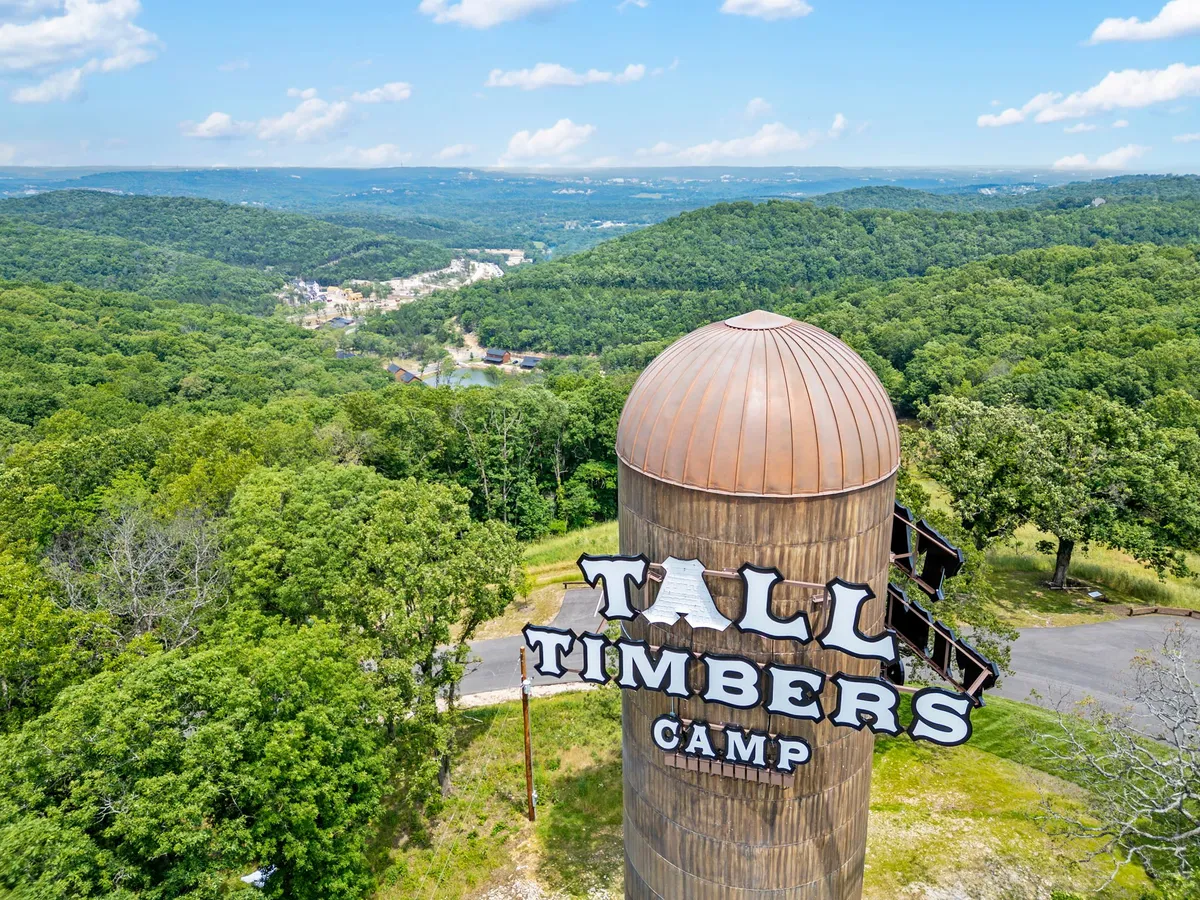 A wide view featuring the Tall Timbers Camp sign set against rolling green hills and a distant mountain range under a clear blue sky. The open space and signage evoke a sense of arrival at an outdoor adventure hub surrounded by nature.