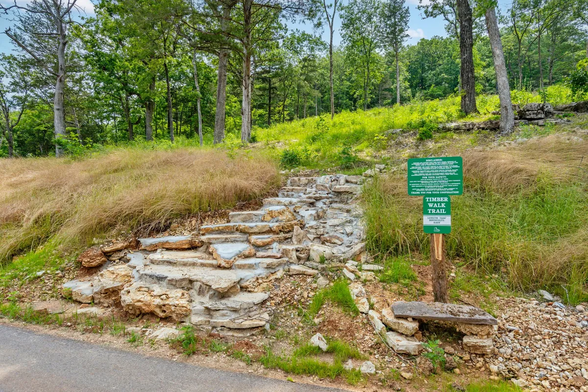 A scenic exterior view of stone steps leading into a lush, green trail, marked by a clear trail sign. The surrounding area is filled with mature trees and vibrant undergrowth, evoking a sense of adventure and immersion in nature. The pathway invites guests to explore the resort's extensive walking trails.