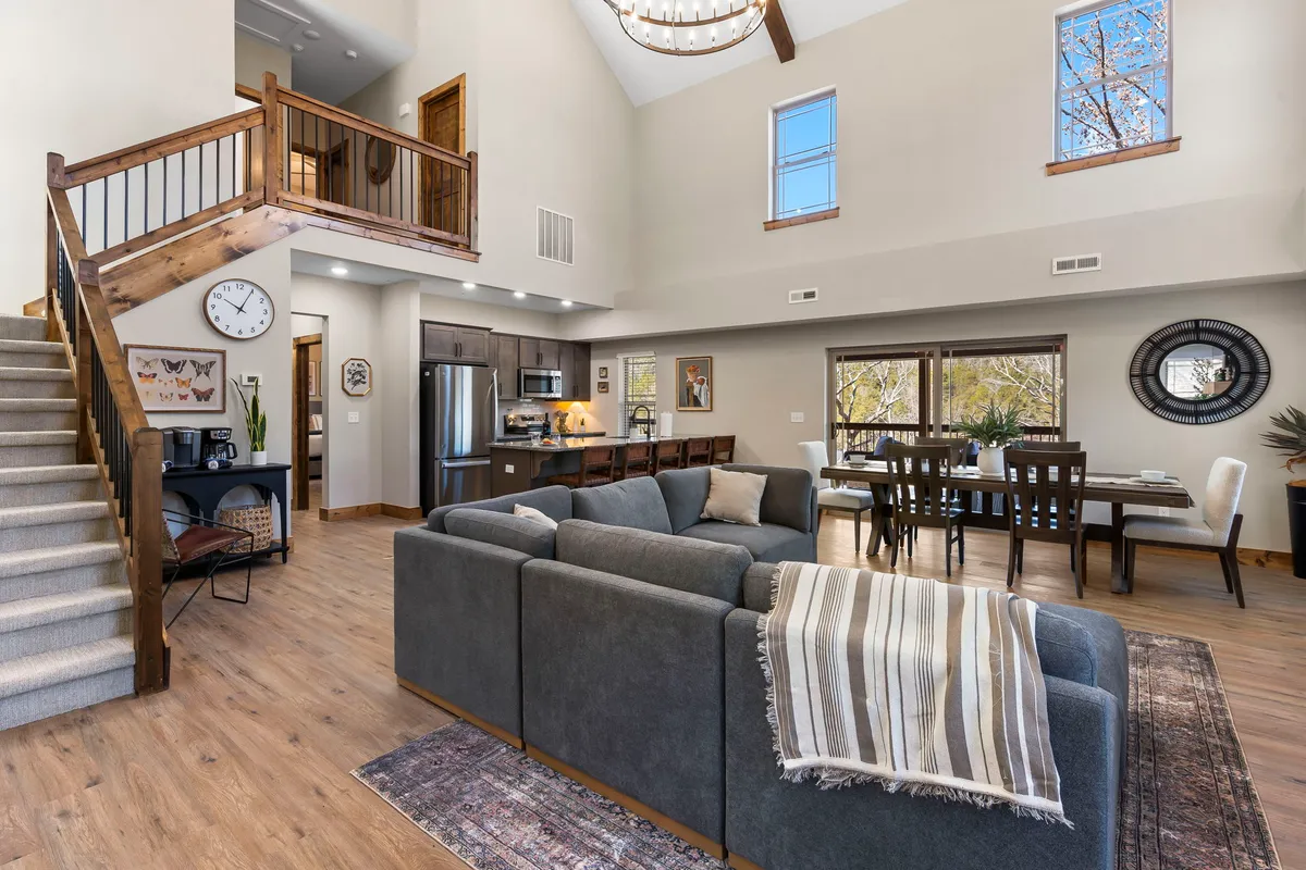 A cozy living and dining room featuring a comfortable sofa, a spacious dining table ideal for meals or game nights, a grand staircase, large windows, and an elegant chandelier. The room is bright, airy, and designed for both relaxation and gathering with loved ones.