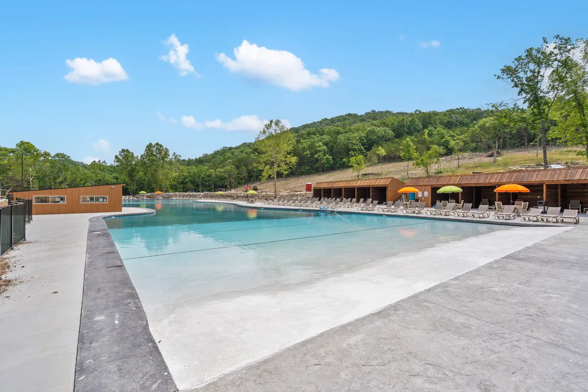A vibrant outdoor pool scene featuring a large swimming pool with colorful umbrellas, lounge chairs, and wooden cabanas, surrounded by natural landscape. The inviting setup is perfect for sunbathing, swimming, and socializing with family or friends.